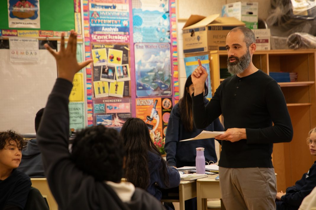 A bearded teacher speaking to his class while holding a paper, with a student in the foreground raising their hand.