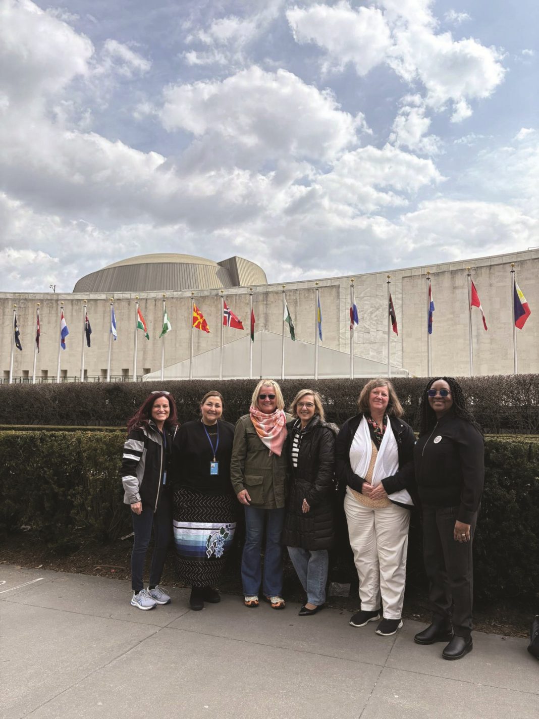 A group of six diverse women posing outdoors in front of the United Nations headquarters with international flags lined up in the background.