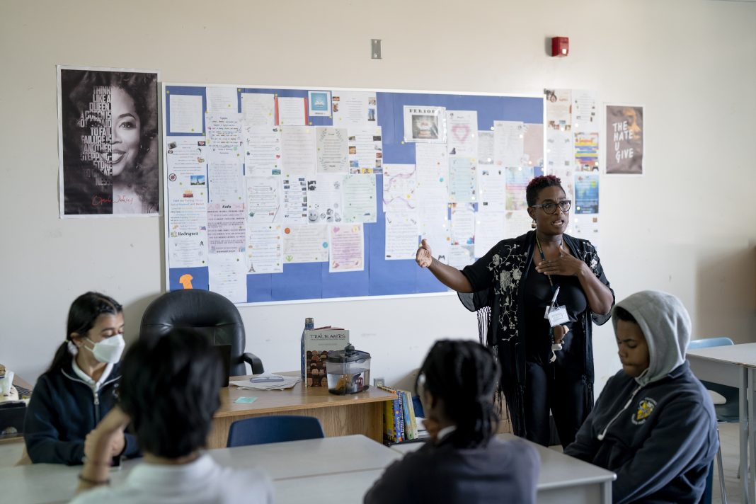 A Black female teacher gestures while speaking to students seated in a classroom with posters on the wall.